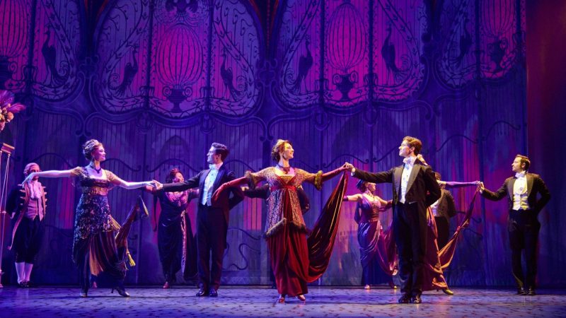  The company of the national tour of "My Fair Lady." Photo by Jeremy Daniel. Cast members wear elegant early 1900s costumes and dance onstage in front of a purple backdrop, onto which the pattern of ornate curtains is projected.