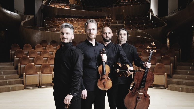  The members of Italian string quartet Quartetto di Cremona stand on stage in an empty theatre, holding their instruments. The quartet, made up of four white men, is wearing all black. The empty brown theatre seats are visible behind them.