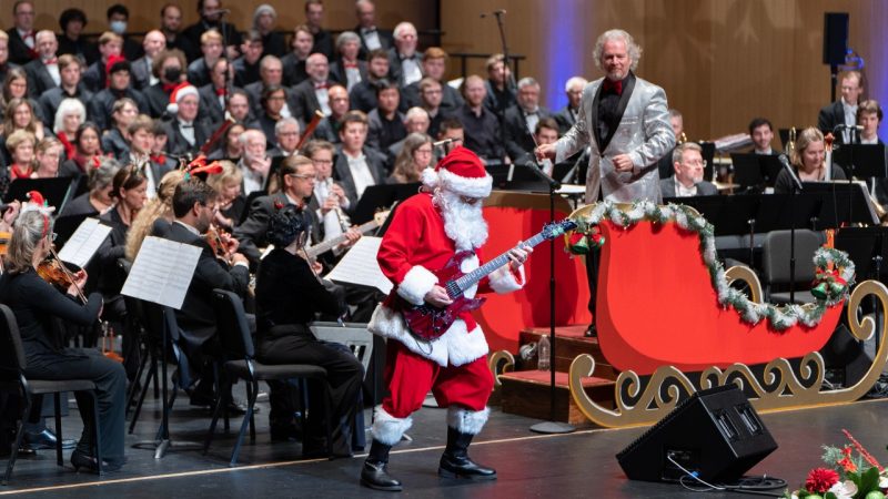 The Roanoke Symphony Orchestra performs its annual "Holiday Pops Spectacular" at the Moss Arts Center. In the foreground, a man in a Santa costume plays a red electric guitar while conductor David Stewart Wiley looks on from a large red sleigh.