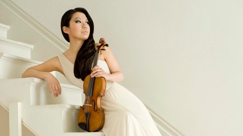 Violinist Sarah Chang, an Asian woman with long dark brown hair, smoky eye makeup, and nude lipstick, holds her violin and reclines on a white staircase in front of a beige wall.
