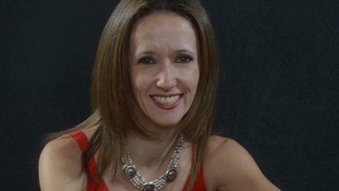  Pianist Sonya Ovrutsky Fensome wears a red tank top and chunky silver necklace and smiles at the camera. She is a white woman with brown eyes and shoulder length, medium brown hair.