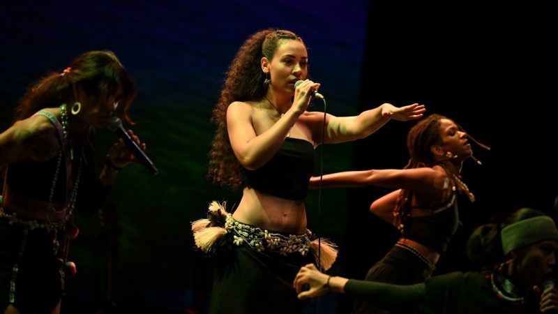 Two young AAPI women wearing traditional dress and accessories perform during "Small Island, Big Song." At left, a Black woman with a white dress, pink and orange scarf tied around her waist, and puka shell jewelry sings and plays a large hand drum. At right, a woman with long brown hair wears a black crop top, black pants, and traditional jewelry and plays a red and white bass guitar.