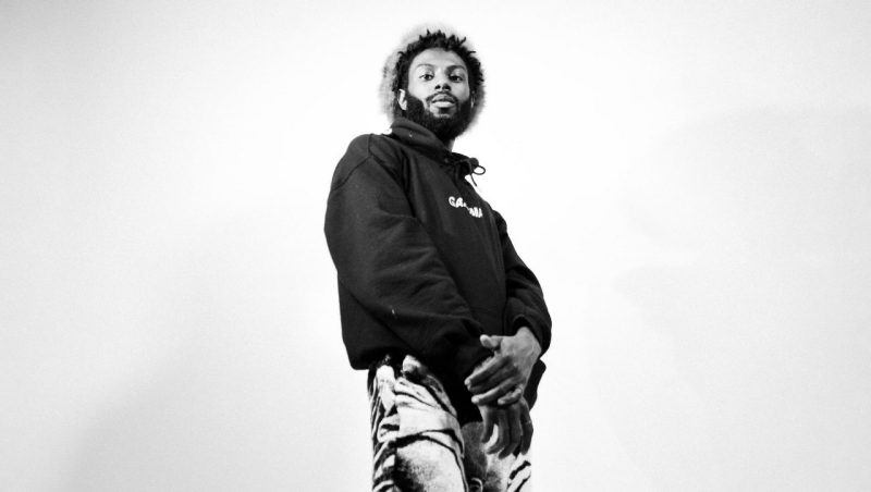  Sonny Miles, a Black man with medium length twists and a full beard, wears a light colored fabric hat, black hoodie, and patterned wide leg pants. He looks down towards the camera, which was close to the floor when the photo was taken, with a neutral expression in this black and white photo.