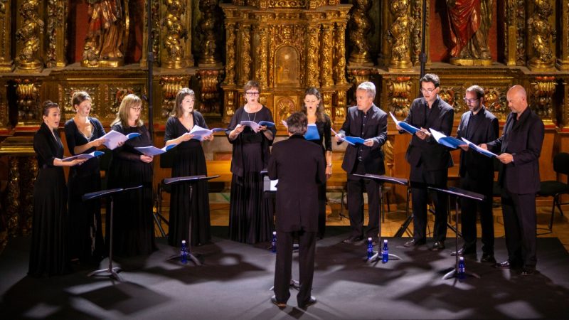 Members of the Tallis Scholars hold music books and sing in front of an ornate church wall. They are wearing all black and are being conducted by director Peter Phillips, who has his back to the camera.