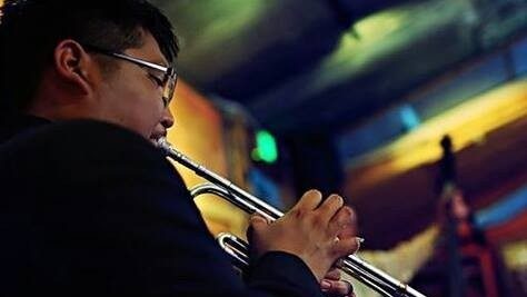  Trumpeter Delbert Anderson, an Indigenous man with dark short hair and glasses, plays his trumpet in what looks like a club.