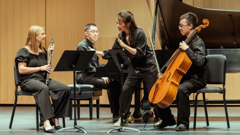Members of Brentano String Quartet lead a master class for students in the School of Performing Arts. At center, a middle aged Asian woman gestures while enthusiastically coaching wind and string student musicians.