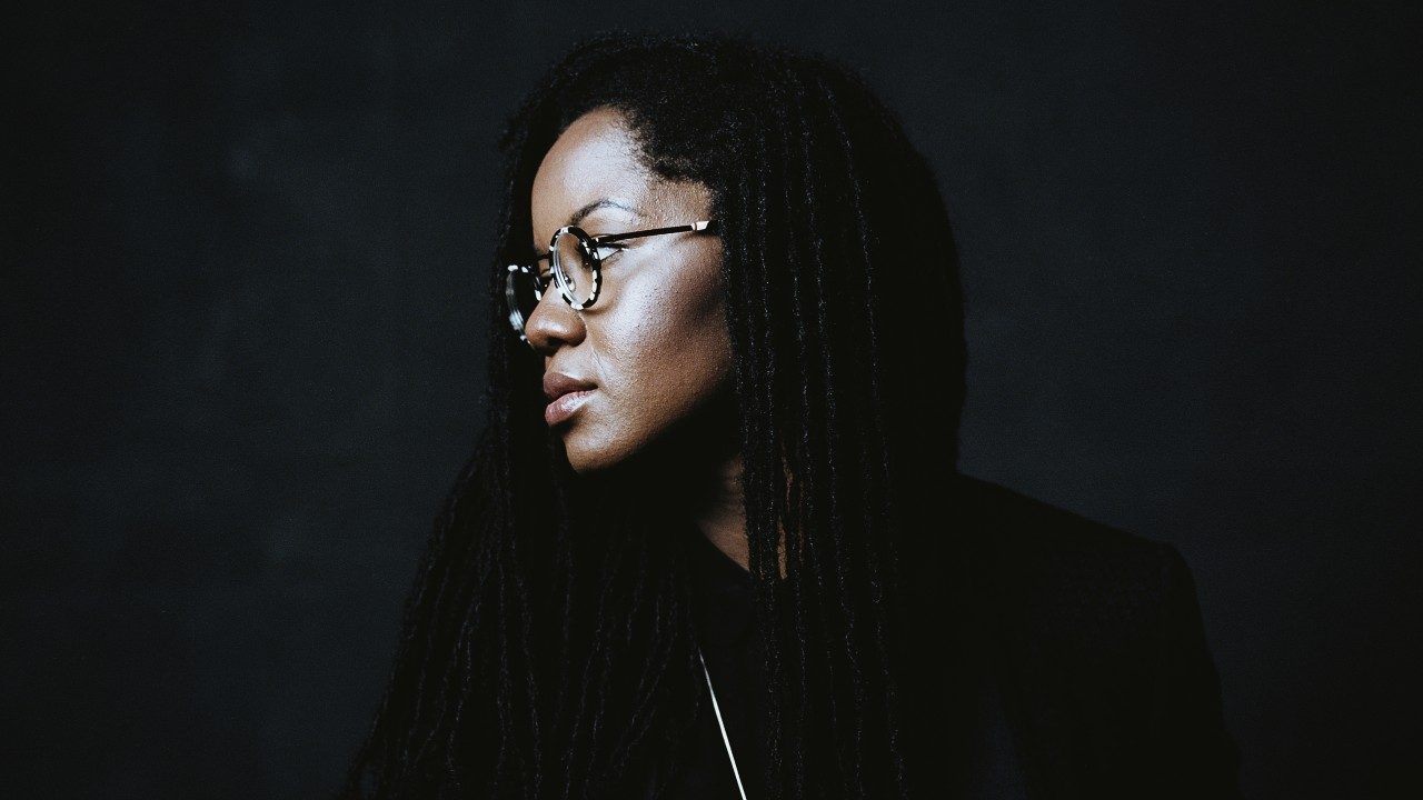  A headshot of Shannon Sea, a Black woman with long braids. Shannon wears a black shirt and round wireframe glasses, and she is looking to the left of the frame.