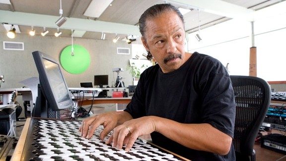 Stephen Taylor, a Black man with a moustache and combed-back hair, sits in a desk chair playing a Wilson Microzone