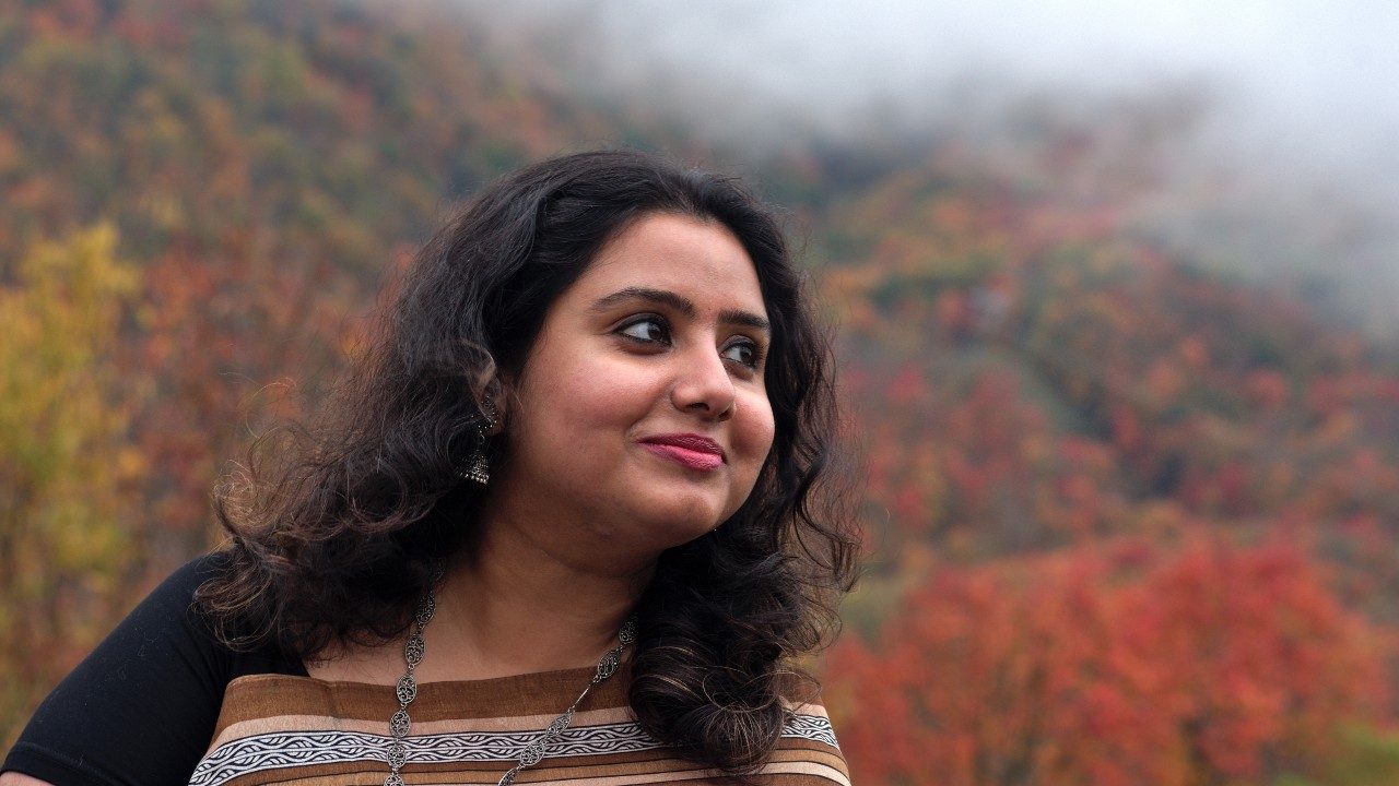  A photograph by Adetoso Adonja of an Indian woman with long dark brown hair and red lipstick wearing a multicolored sari. She looks to her left and smiles. Behind her rises a mountain covered with trees changing colors in the fall. At the top of the image is a swath of fog.