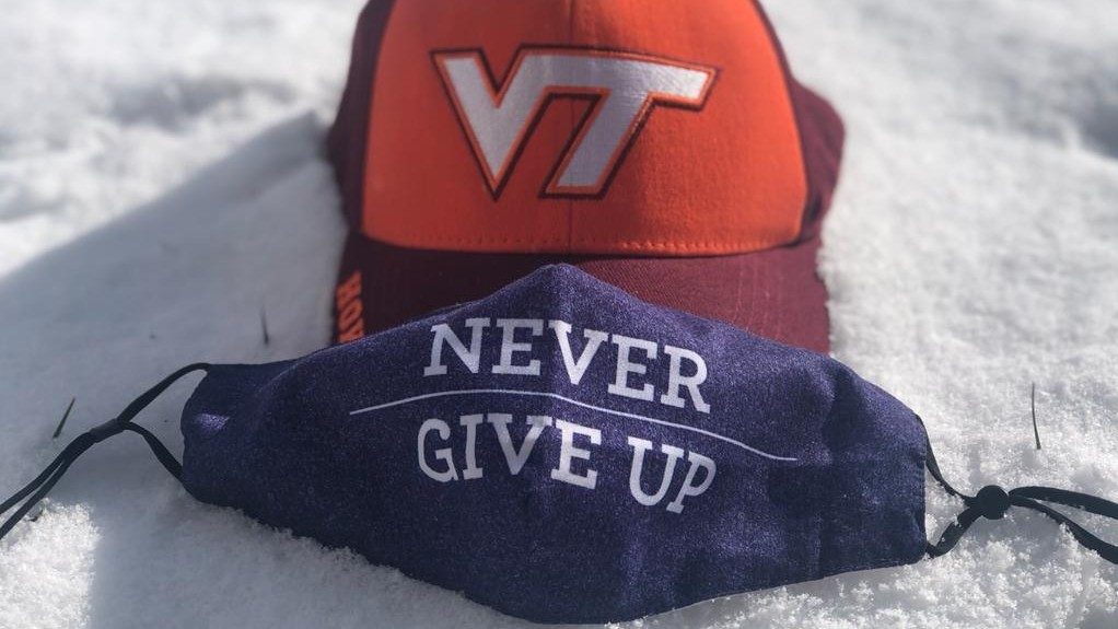  A photograph by Kamla Al Amri of a VT baseball cap with a maroon bill and orange top, onto which the Virginia Tech athletics logo is printed. Overlaid on the bill of the hat is a navy face mask that reads "Never Give Up." Both are sitting on top of snow in the sunlight.