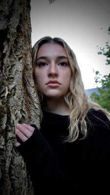  A photograph by Abigail Alisesky of a young woman with long blonde hair leaning against a tree. She is wearing a dark long sleeved sweater and looking at the camera. Behind her are green leaves and the ridge of a mountain.
