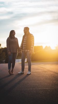  A photograph by Travis Carr of a young white couple holding hands and looking at each other. They stand in an asphalt parking lot with the sunrise behind them. There is light cloud cover. The woman, on the left, wears a blush pink sweater, jeans, and tan booties, and has shoulder length light brown hair. The man, on the right, wears glasses, a dark vest, a plaid button down flannel shirt, jeans, and sneakers.