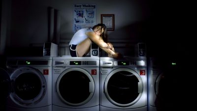  A photograph by Kirsten Doane of a white woman with long dark brown hair in a white shirt and dark athletic shorts sitting on top of two washing machines in a laundromat. The room is dark except for natural light coming in through a window. The woman sits with her knees bent at a 90 degree angle, with her arms wrapped around her legs and her head resting on top of her knees. She looks towards the camera.