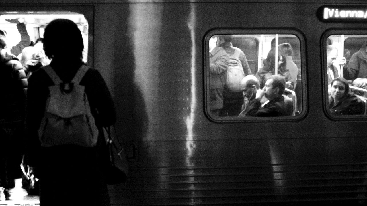  A black and white photograph by Eugene Lim of a metro train headed to Vienna/Fairfax. The image is taken from the train platform looking towards the car. The door is open and people board; in the window, you can see passengers sitting and standing.