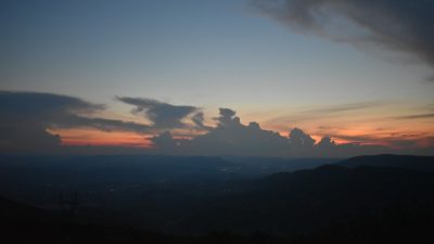  A photograph by Reaz Mahmood of a sunset behind a mountain range. The mountains are towards the bottom of the frame and are dark. Just above the mountains is a strip of bright pink obscured in part by dark clouds. The sky above fades to a dark blue.