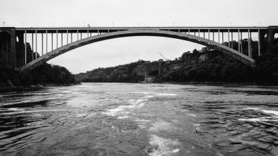  A black and white photograph by Rupabali Samanta of an arched metal bridge over a river with thick trees on either side of the river. The photo was taken from the back of a boat, and the wake is visible in the water.