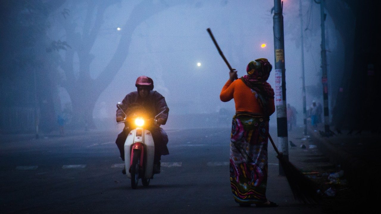  A photograph by Md Hasan Shahriar of a foggy morning on a street in Dhaka.  On the left, a person rides by on a moped with the headlight on; on the right, a person sweeps. In the back, you can make out the form of a tree through the fog.