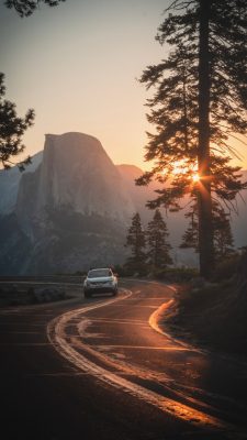  A photograph by Sean Stroud of a gently winding mountain road. A small white SUV is traveling in the right lane towards the camera. Tall pine trees are scattered along the sides of the road. In the background is a tall mountain ridge, and the sun peeks above the mountains, either at sunrise or sunset, and filters through the boughs of a large pine tree. The sky is a clear light pinky blue.