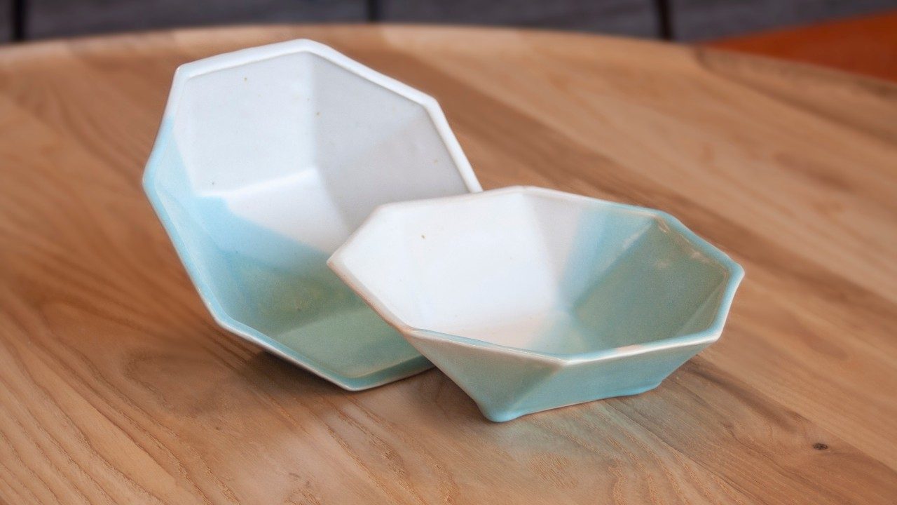 An octagonal ceramic form, painted or glazed half white and half light blue. The two pieces sit with the insides facing out on a wooden table.