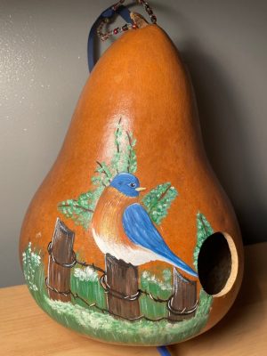  A gourd turned into a birdhouse painted with acrylic paint by Alexis Lawson. The gourd is painted a burnt umber base, and on top of that is a blue bird looking towards the birdhouse opening, with a reddish brown chest and white feathers towards where its feet would be. It is perched on a brown picket fence connected with metal wire. Behind the fence and the bird is green grass with some white flowers and a small green tree.