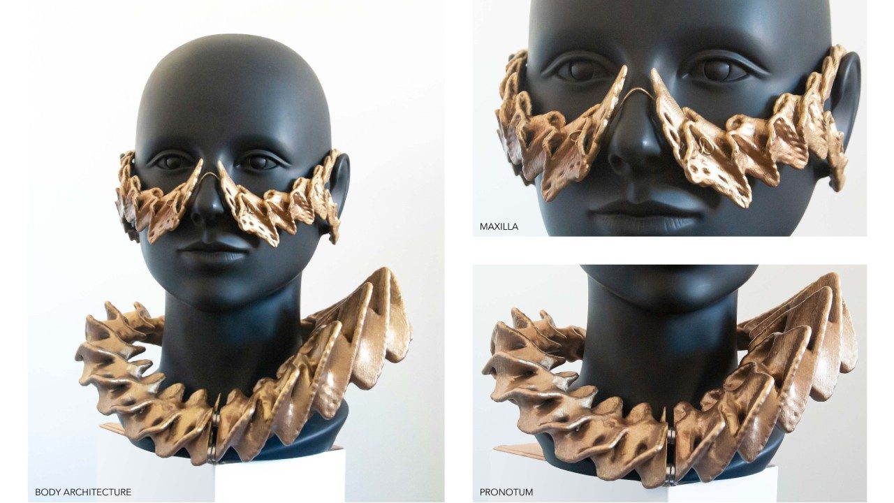  RJ Weaver's laser scanned computational digital design and 3D printed body armor. A gold-colored 3D-printed necklace and face armor are modeled on a black mannequin head. The pieces are sculptural and ridged. The necklace sits away from the neck and comes up on the right and left sides, almost in peaks. The face piece nestles over the nose using a bridge like what is used in glasses, and hooks over the ears.