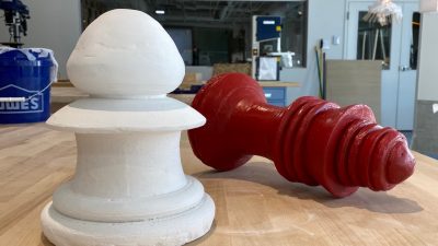  Two fabriacted oversized plaster chess pieces on a wooden table in the studio where they were created.  The one in the foreground is shorter and white with a rounded top. The one in the background is taller, red, and knocked on its side, as if defeated during play.