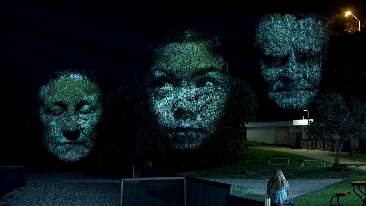  An example of Craig Walsh's "Monuments," enormous night-time projections that transform trees into sculptural monuments. Three faces are being projected onto trees, and a white woman with long blonde hair and a white shirt sits on a bench to watch them.