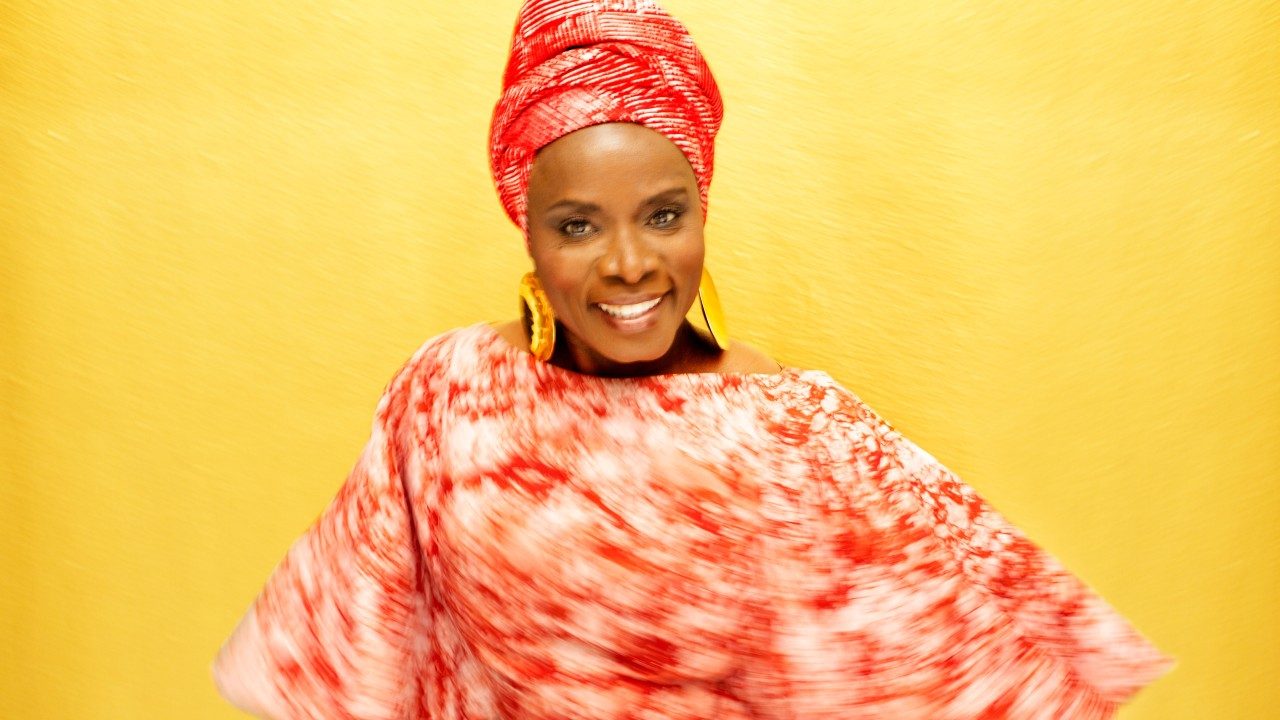  Singer Angélique Kidjo twirls in a red and white patterned dress, red hair wrap, and gold earrings in front of a yellow background.