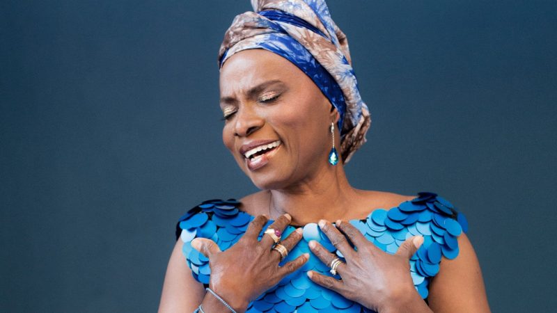  Singer Angélique Kidjo wears a blue dress with large flat sequins, blue teardrop earrings, and a blue and brown hair wrap. She holds her hands towards her collar bone and sings with her eyes closed and her face angled away from the camera.