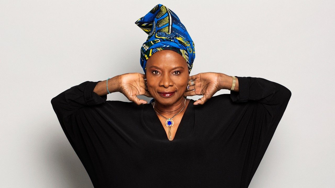  Singer Angélique Kidjo wears a black sleeved dress, blue necklace, and a blue and yellow patterned hair wrap. She holds her hands towards the back of her neck with her elbows pointed in opposite directions and smiles towards the camera.