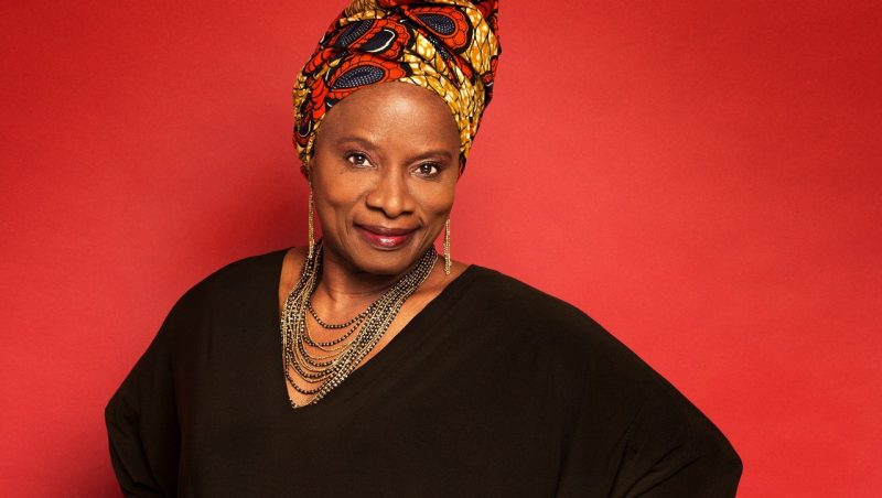  Singer Angélique Kidjo wears a black sleeved dress, a gold multistranded necklace and earrings, and red and gold patterned hair wrap and stands in front of a red background.