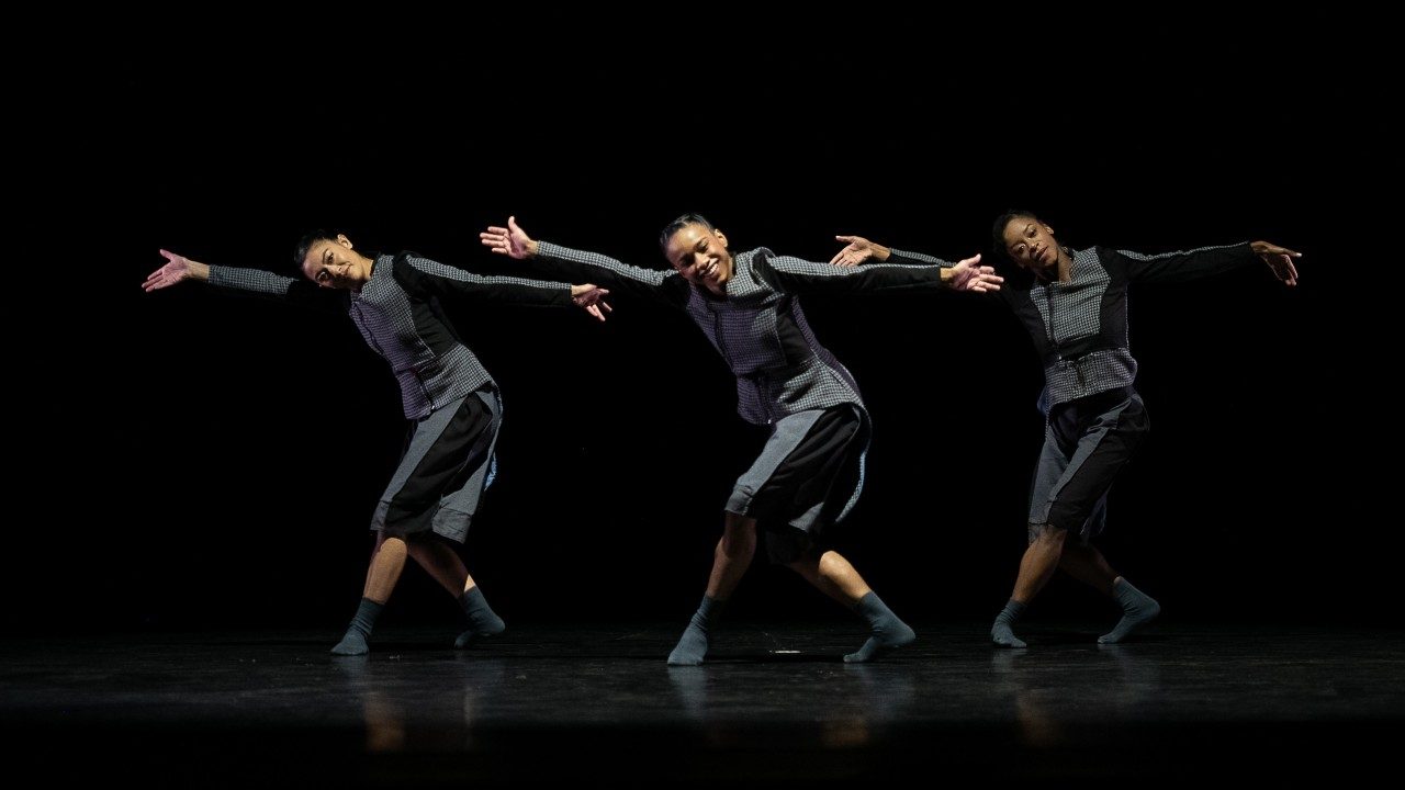  Three dancers from Ballet Hispanico perform on stage in matching grey costumes. All three have their arms extended to both sides, and are stepping to the right, with their left leg crossed in front of their right legs.