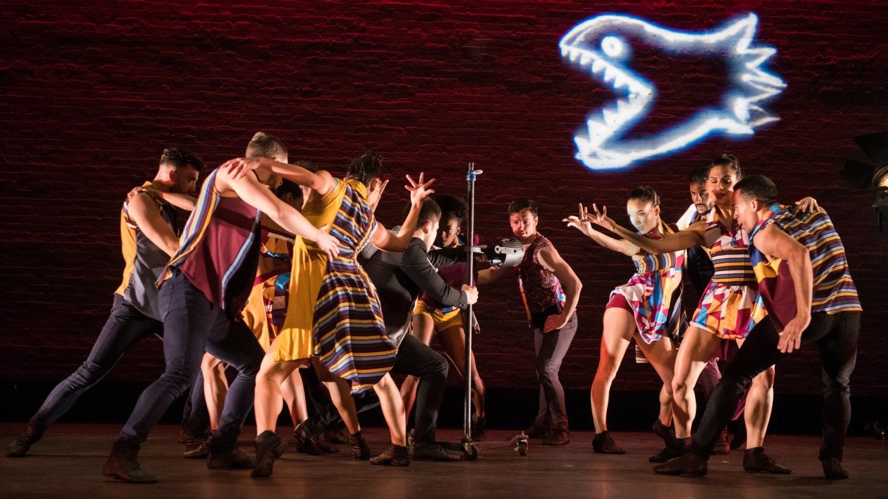  The dancers of Ballet Hispánico on stage in multicolored costumes dance around a light stand with a projection of a white fish with teeth on a brick wall behind them.