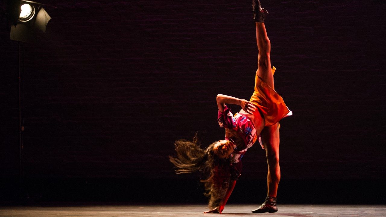  A dancer from Ballet Hispanico dances on stage. She is standing on one foot, the other extended up into a full split, and she reaches down to the floor with her right hand.