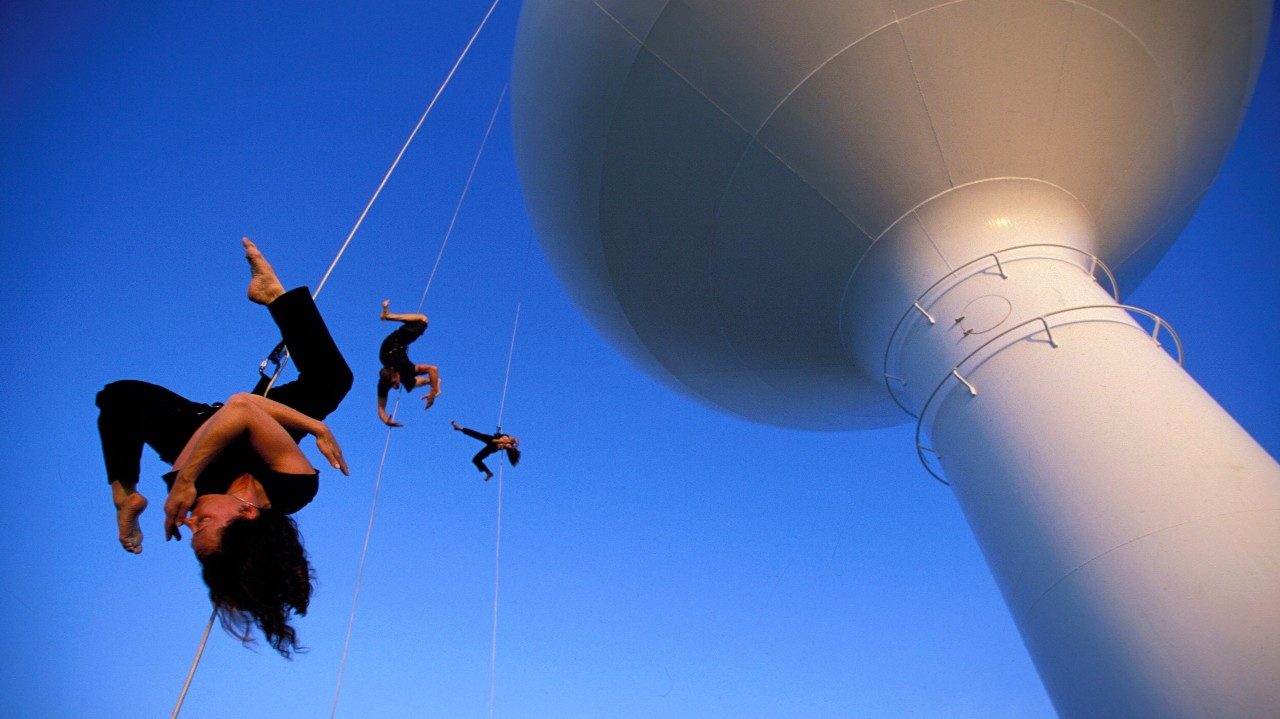  The dancers of BANDALOOP dance while suspended from the side of a water tower. Three dances in dark clothes hang from climbing equipment.  The background is a clear blue, and the water tower is white. 