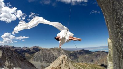 A female dancer from BANDALOOP dances against a rock face in Yosemite, suspended by climbing equipment. The dancer wears white and is draped in a long flowing white fabric. The sky is a perfect deep blue with a few patchy pure white clouds. In the far background is a mountain range and a lake.