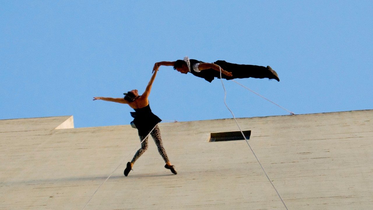  Two dancers from BANDALOOP dance against the side of an exterior wall, suspended by climbing equipment. The female dancer is on tiptoes, head arching back, and arms extended, and the male dancer is completely parallel to the wall and touching only the female dancer's outstretched hand.