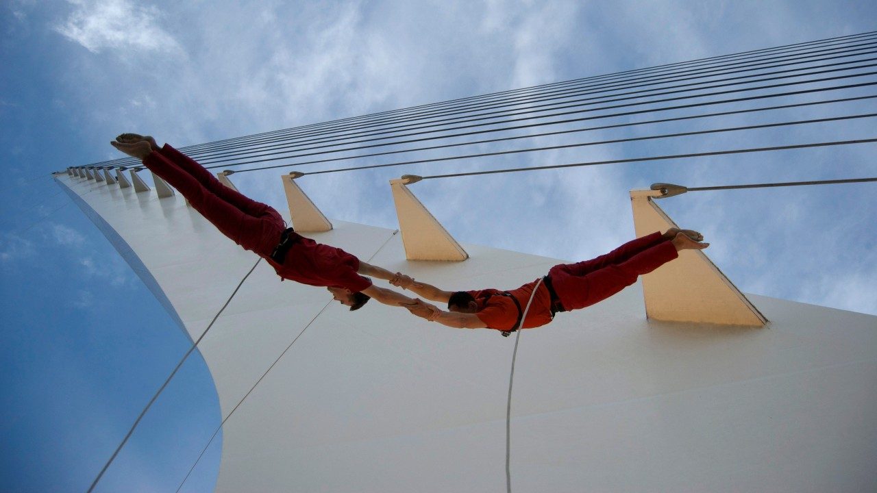  Two male dancers from BANDALOOP dance against the side of Sundial Bridge wearing orange shirts and pants. They are suspended in mid air by climbing equipment. Behind them are 14 metal support cables in horizontal lines. 