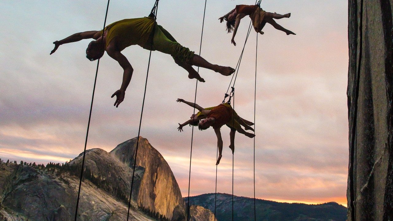  Dancers from BANDALOOP dance against Mount Watkins in Yosemite National Park in California. The dancers are suspended in mid air by climbing equipment. Behind them is a mountain range awash in pink light; the clouds are cotton candy pink, giving way to light blue sky. Just above the mountain is a strip of brilliant orange.