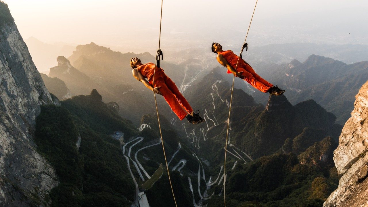  Dancers from BANDALOOP dance against Tianmen Mountain in China. The dancers wear orange shirts and pants and jump directly away from the mountain, arms at their sides, suspended in mid air by climbing equipment. Far below them is a winding mountain road. In the background is a mountain range awash in soft light.
