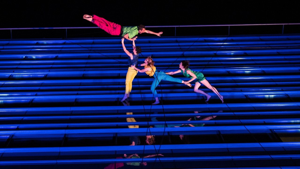  Four dancers from BANDALOOP dance against the exterior wall of a building in Budapest, suspended by climbing equipment. The building is lit blue and covered in windows. The dancers are wearing brightly colored costumes