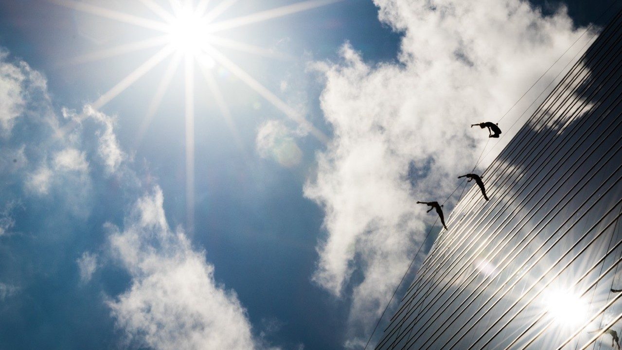  Three BANDALOOP dancers against the exterior wall of IMAX Dallas, suspended in mid air by climbing equipment. The image has been shot from below them on the ground.. The sun is shining brightly and reflecting off the glass of the building;  a few clouds are in the sky.