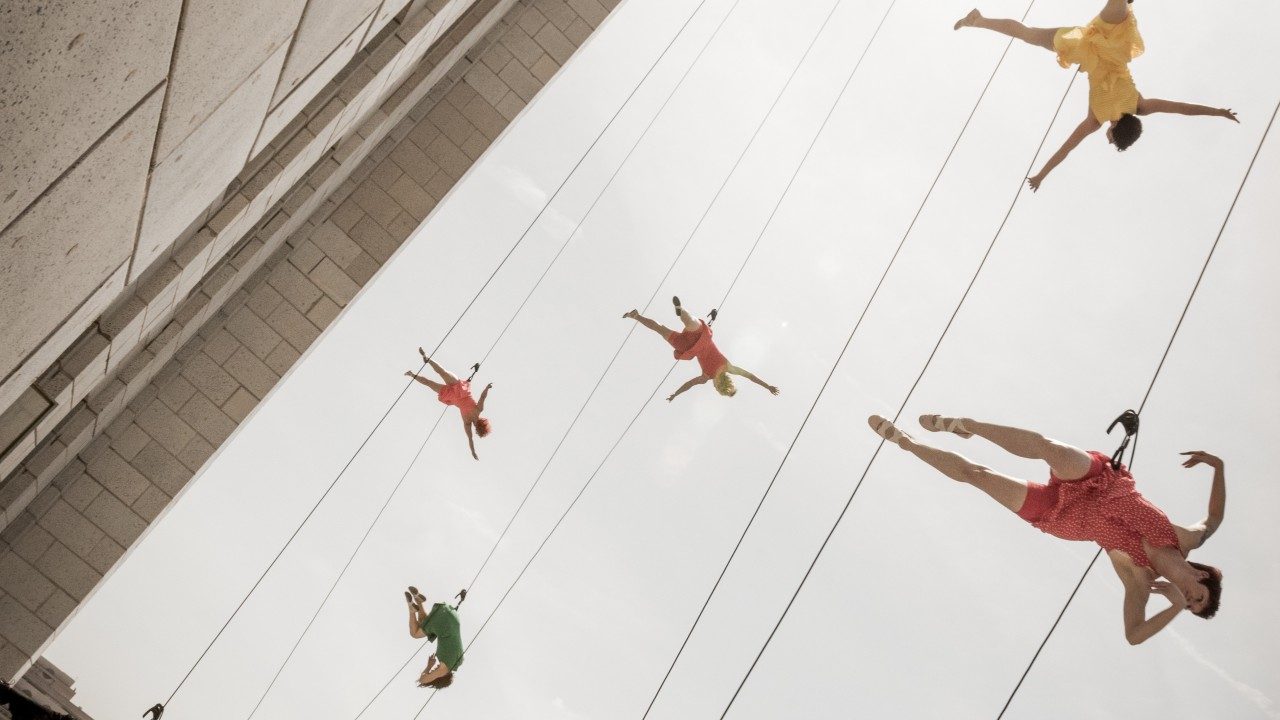  Five BANDALOOP dancers in various colors (green, orange, pink, and yellow) dance against an exterior wall in Los Angeles, suspended in mid air by climbing equipment. The photo has been taken from below.