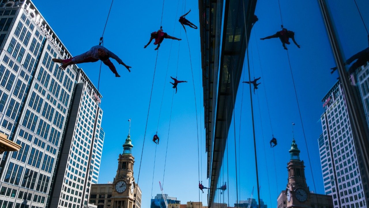  Five BANDALOOP dancers in Sydney, Australia, dance against the mirror glass of an exterior building wall, suspended in mid air by climbing equipment. The sky is a crystal clear blue. Behind the dancers are various city buildings and a clocktower.