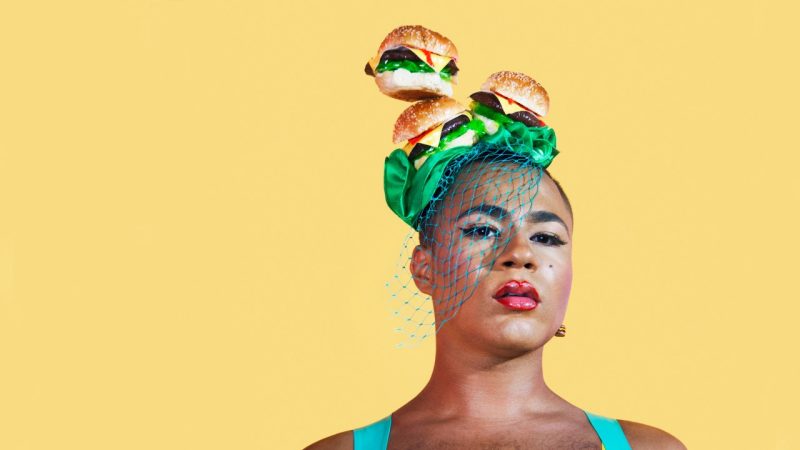 Travis Alabanza wears a turquoise tank top, flawless makeup with a pink lip, and a hat made of three burgers, green ribbon, and teal netting