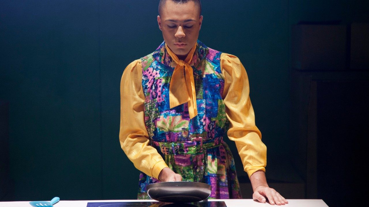  Travis Alabanza wears a yellow-sleeved dress with purple, blue, green, and orange florals, and their hair is in a top knot. They hold a skillet over a glass cooktop, with a turquoise spatula nearby.