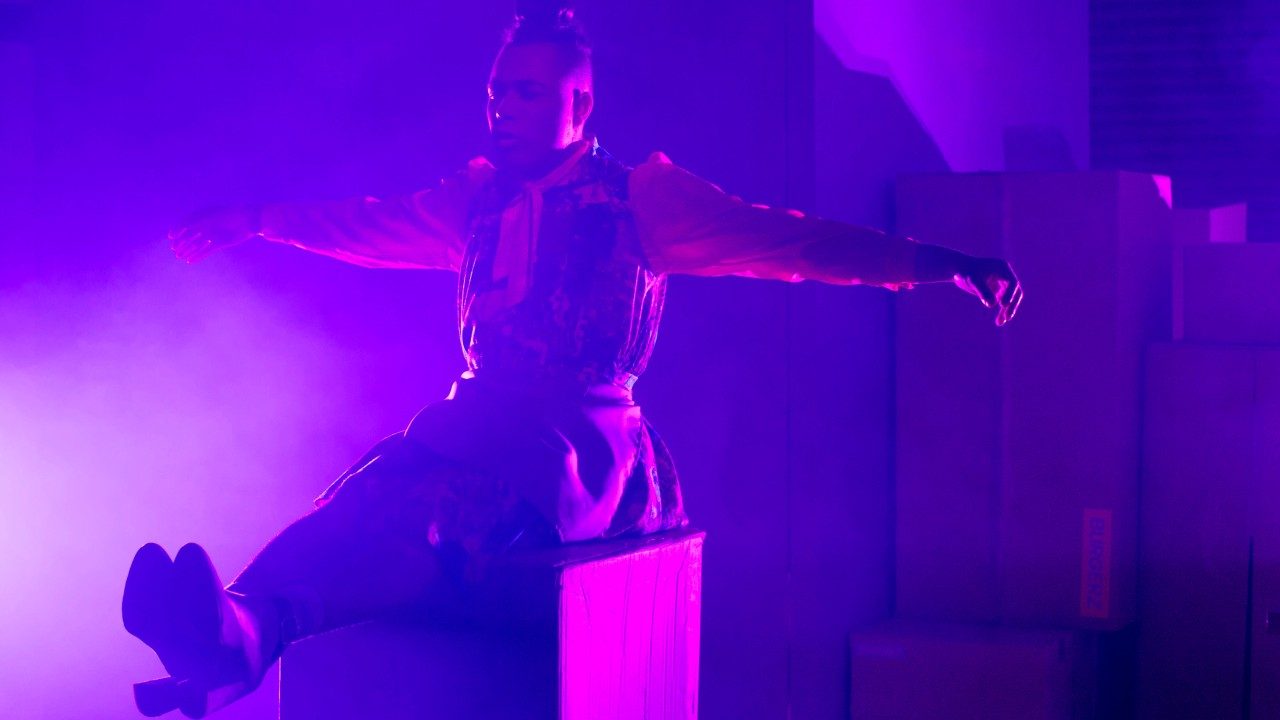  Travis Alabanza sits on a box on stage during their production of "Burgerz." They wear a multicolored dress, a top knot, and heels, and they sit with their legs together and extended straight out in front of them, and their arms are extended to either side. The stage is lit in a purple/pink light.