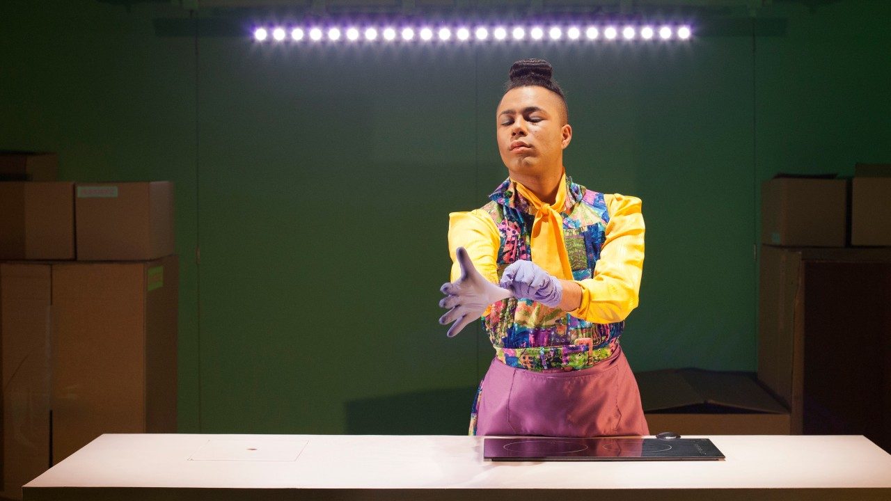  Travis Alabanza wears a multicolored dress with yellow sleeves and a yellow neck bow. Their hair is in a top knot and they have a pink apron tied around their waist. They are pulling on lavender latex gloves and stand in front of a counter with a cooktop. Behind them is a strip of lights, lots of cardboard boxes, and a green wall.