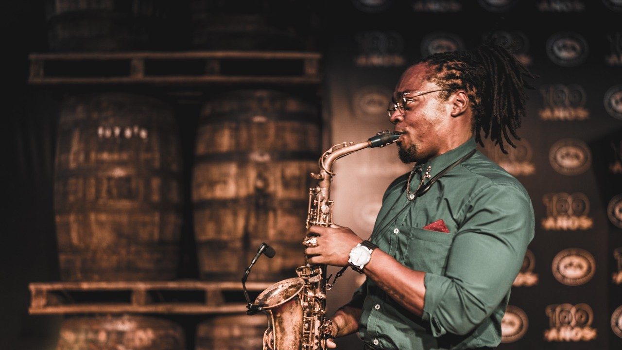  A Black man with braids pulled back into a pony tail plays a saxophone. He wears an army green button down shirt with a red pocket square, a black watch, and thin framed glasses.