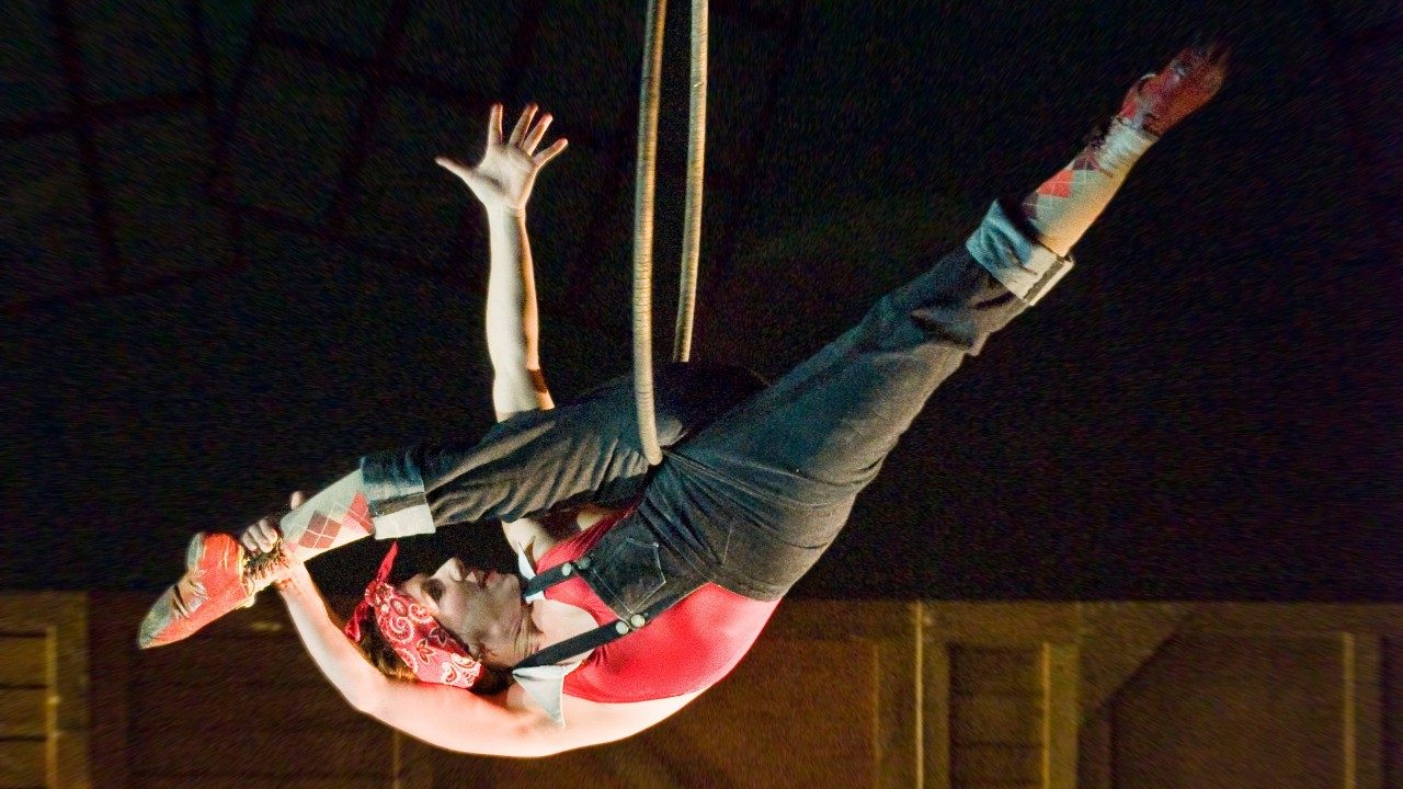  A woman in black overalls, a red shirt, a red bandana, and red argyle socks dangles from a trapeze hoop.